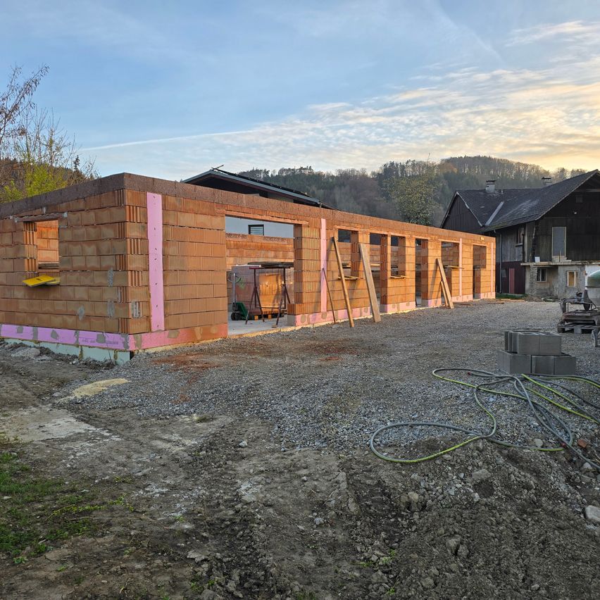 Eine Baustelle mit einem Haus im Bau, umgeben von Kies und mit unfertigen Fenstern und Türen. Im Hintergrund befinden sich zwei weitere Häuser.
