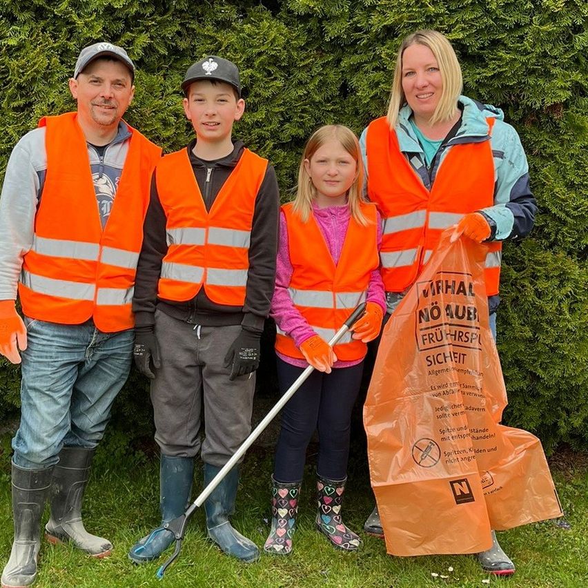 Vier Personen stehen vor einer Hecke und tragen orange Sicherheitswesten. Ein Mann, eine Frau und zwei Kinder posieren für ein Foto. Das Mädchen hält einen Plastikbeutel.