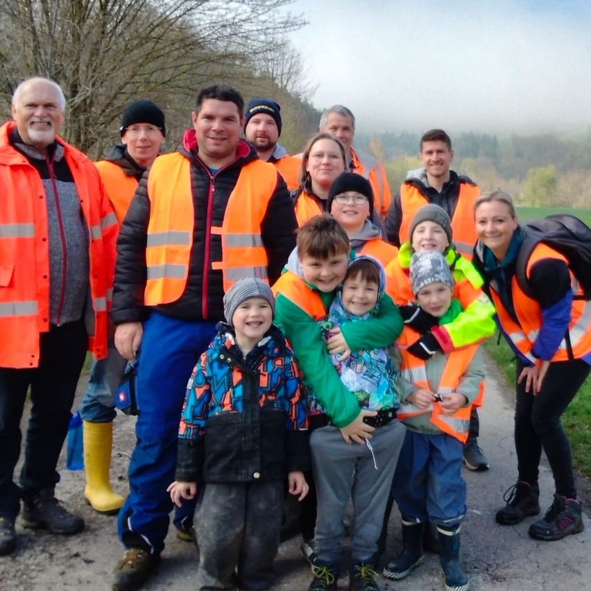 Eine Gruppe von Menschen, einige in Sicherheitswesten, posiert für ein Foto auf einem Kiesweg. Hinter ihnen trägt eine Frau einen Rucksack. Bäume und Berge sind im Hintergrund.