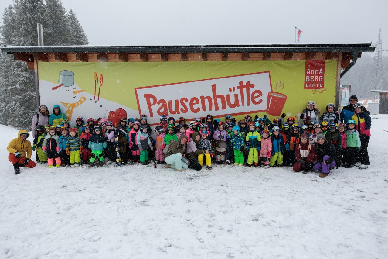 Eine Gruppe von Kindern in Winterkleidung posiert vor einem Banner, das eine Pausenhütte bewirbt.