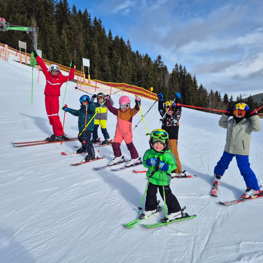 Eine Gruppe von Kindern und einem erwachsenen Skilehrer fährt eine verschneite Piste hinunter, alle tragen Helme und halten Skistöcke. Die Kinder sind in bunten Ski-Outfits gekleidet.