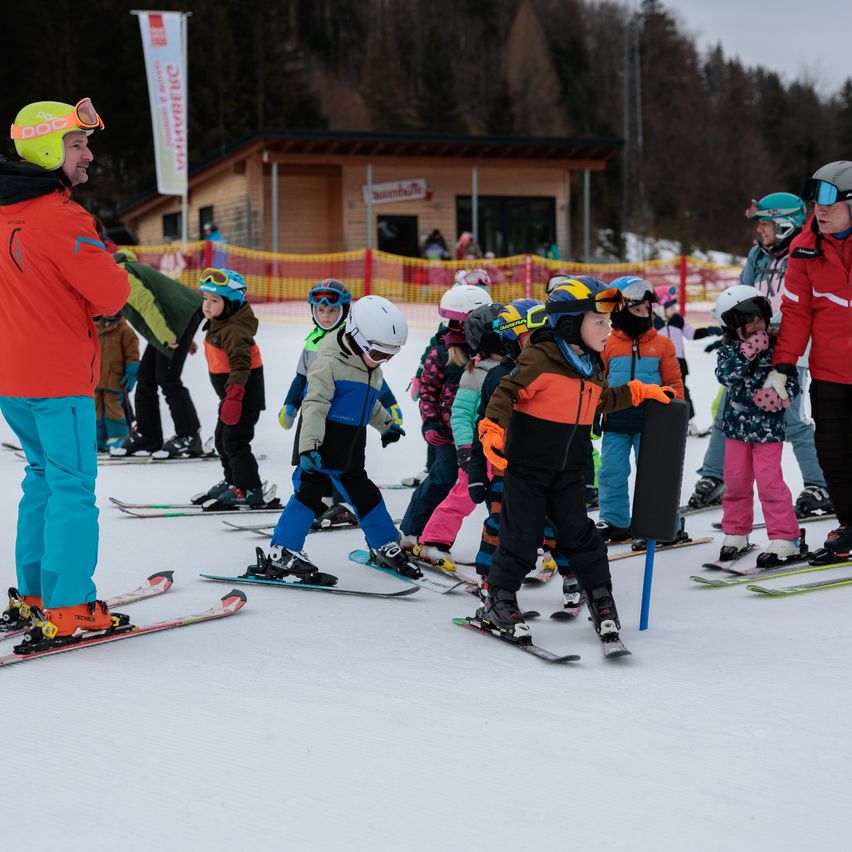 Eine Gruppe von Kindern lernt mit ihren Lehrern auf einem verschneiten Feld Skifahren, mit einem kleinen Gebäude und einem Zaun im Hintergrund.