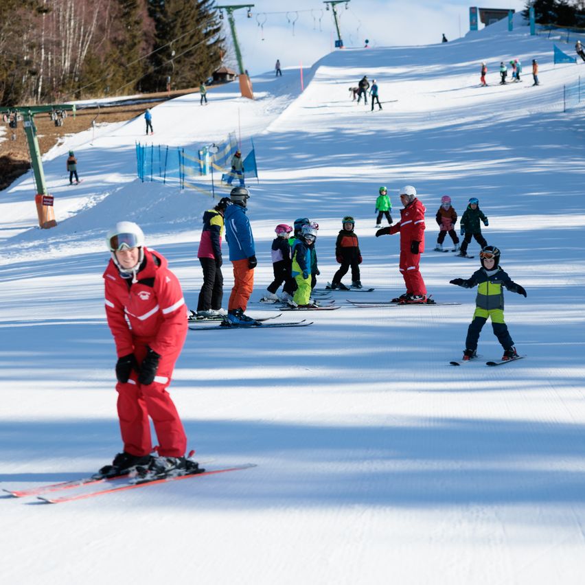 Ein Skilehrer unterrichtet Kinder auf einem verschneiten Hang, während andere im Hintergrund Ski fahren.