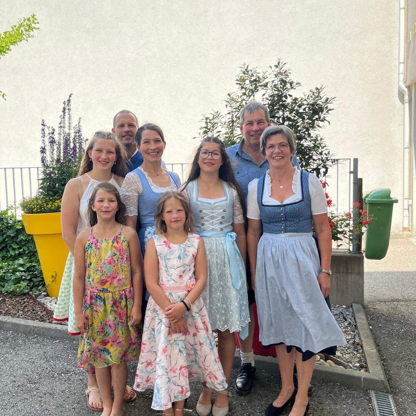 Eine Familie mit acht Personen, darunter zwei Erwachsene und sechs Kinder, posiert für ein Foto vor einem Gebäude mit einem gelben Pflanzgefäß und einem grünen Mülleimer.