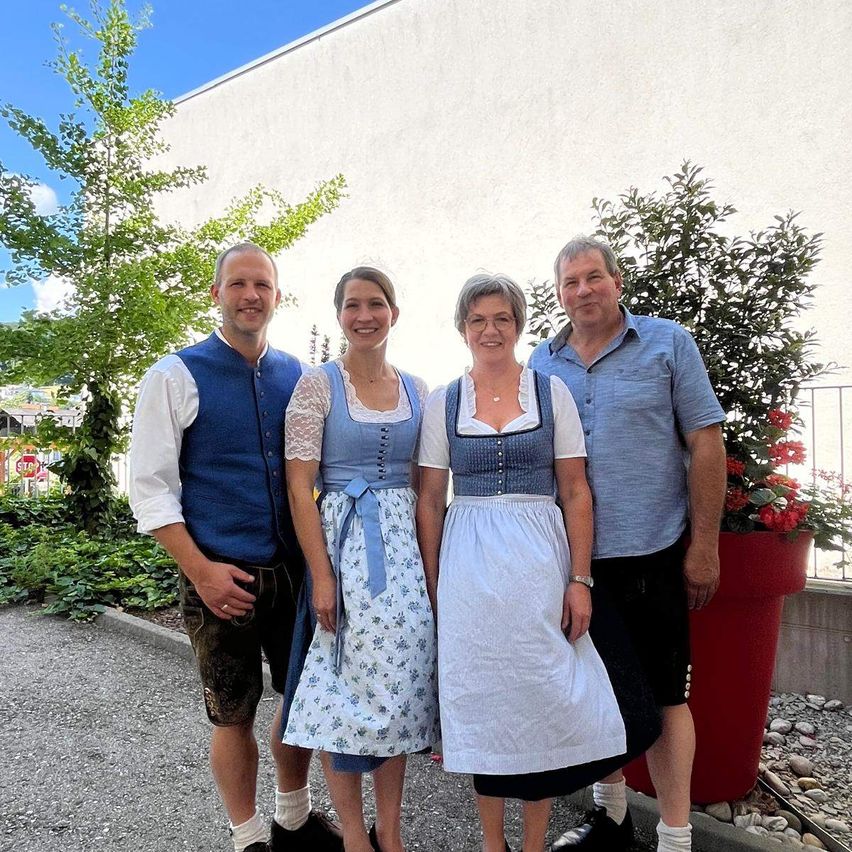 Vier Menschen stehen zusammen vor einem Gebäude, zwei Männer und zwei Frauen, alle lächeln. Sie sind in traditioneller deutscher Kleidung gekleidet. Hinter ihnen stehen Pflanzen und ein Stoppschild.