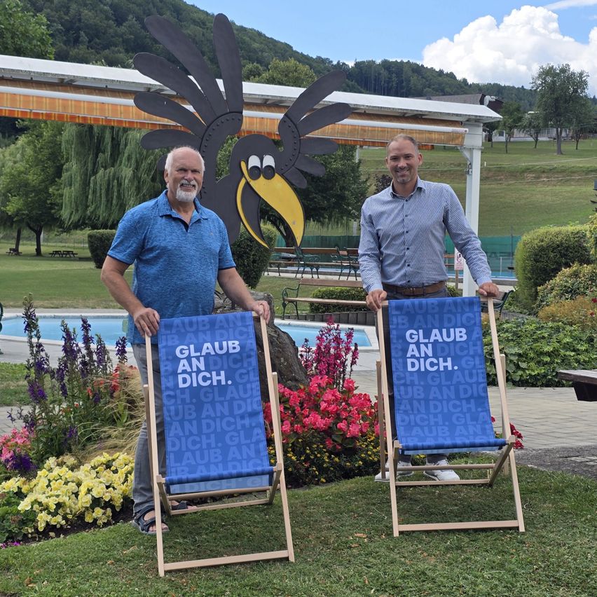 Zwei Männer stehen nebeneinander vor einem großen blauen Schild mit dem Text 'Glaub an dich' in einem Garten mit Blumen und einem Pool im Hintergrund.