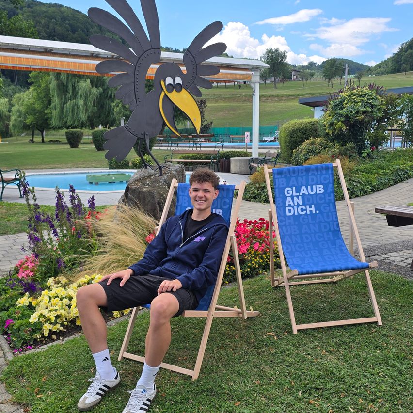 Ein junger Mann sitzt auf einem blauen Liegestuhl in einem Garten mit einer großen Krähenstatue im Hintergrund.