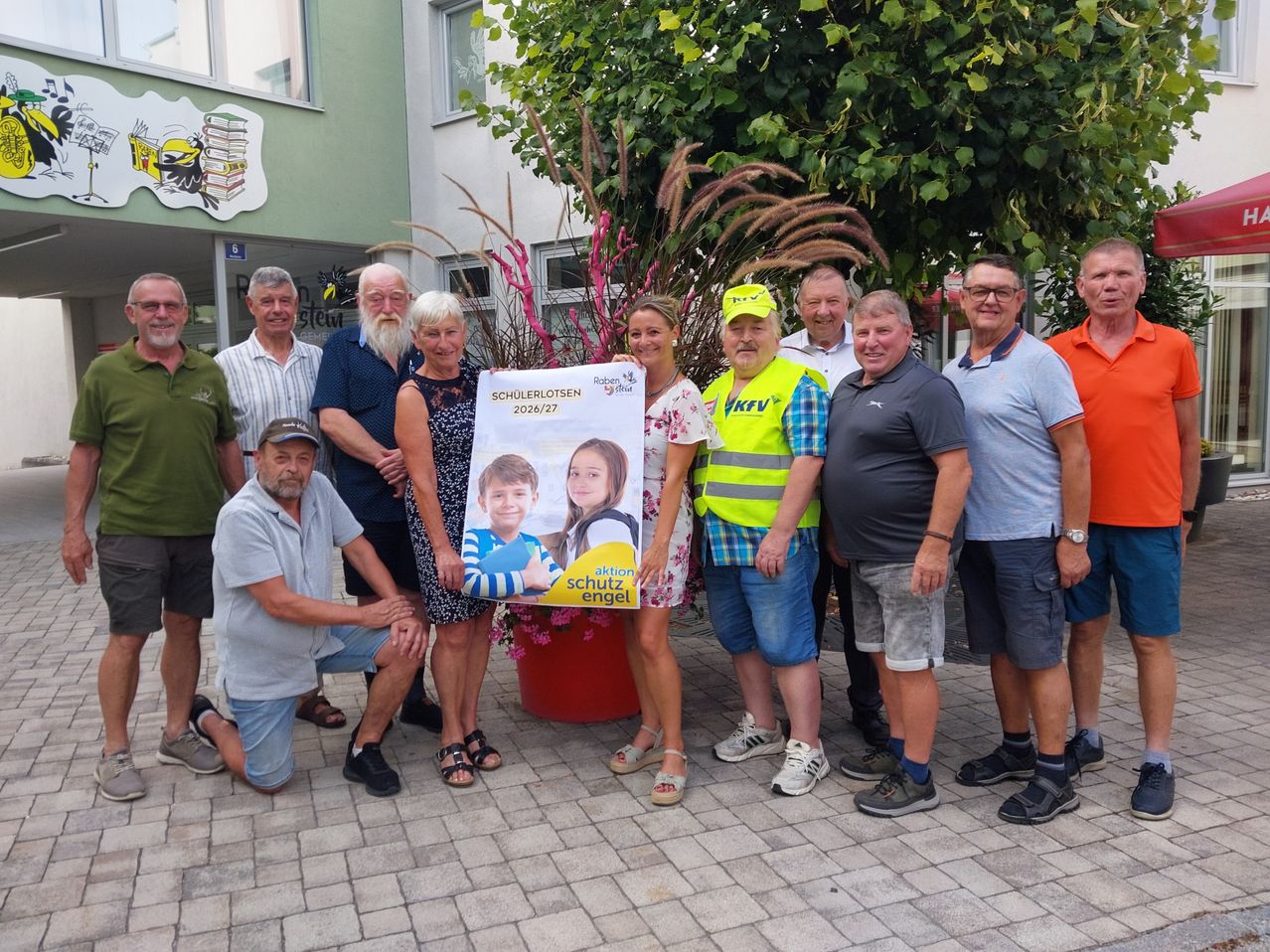 Eine Gruppe Erwachsener steht draußen, einer hält ein Poster mit Fotos von zwei Kindern und dem Text 'Schulerloten 2026/27'.