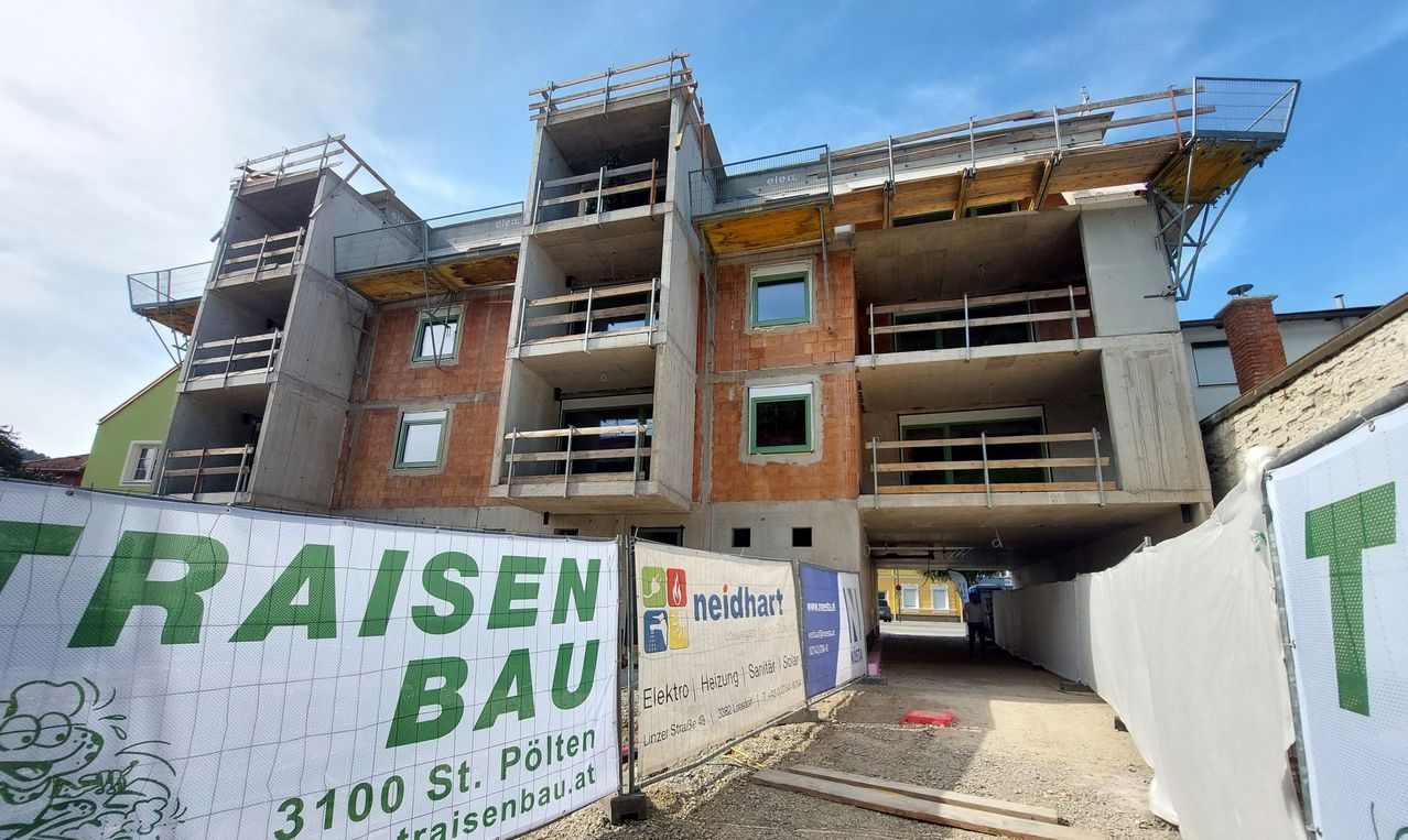 Baustelle mit einem mehrstöckigen Gebäude im Bau, mit Balkonen und Gerüsten. Ein großes Schild auf der linken Seite steht 'AISEN BAU'. Eine Person geht im Hintergrund.