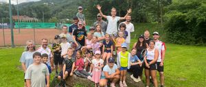 Eine Gruppe von Erwachsenen und Kindern posiert für ein Foto auf einer Rasenfläche mit einem Felsen, Tennisschlägern, einem Tennisplatz und einem Hügel im Hintergrund.