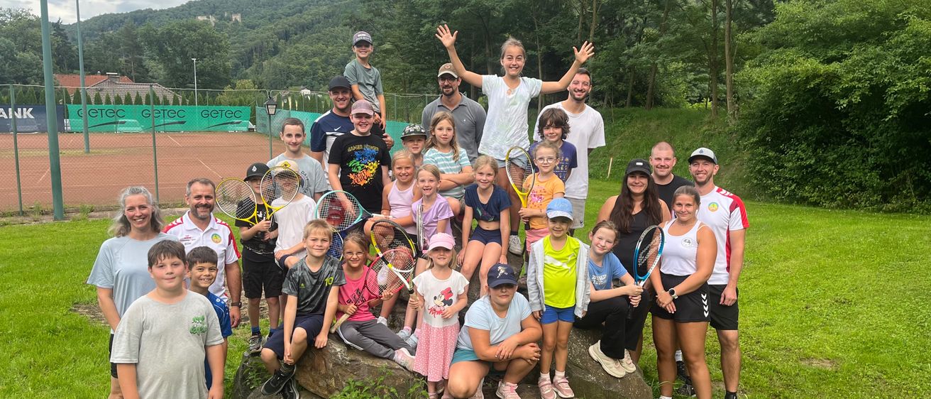 Eine Gruppe von Erwachsenen und Kindern posiert für ein Foto auf einer Rasenfläche mit einem Felsen, Tennisschlägern, einem Tennisplatz und einem Hügel im Hintergrund.