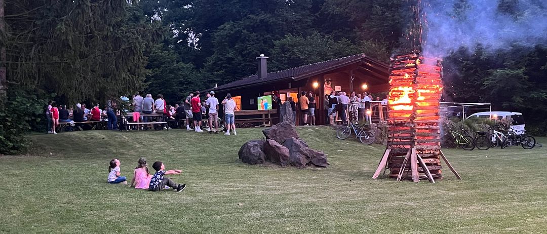Ein Lagerfeuer brennt hell auf einer Wiese, während sich Menschen versammeln, einige sitzen und andere stehen um ein Holzgebäude. In der Nähe sind Fahrräder geparkt.