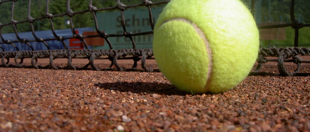 Ein Tennisball liegt auf einem Sandplatz, teilweise im Schatten. Das Spielfeld ist von einem Netz umrandet. Bäume und ein Gebäude sind im Hintergrund zu sehen.