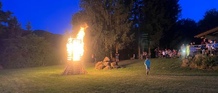 A bonfire burns brightly at night in a grassy field. People are watching from the sides. A boy runs towards the fire. Trees and rocks surround the area.