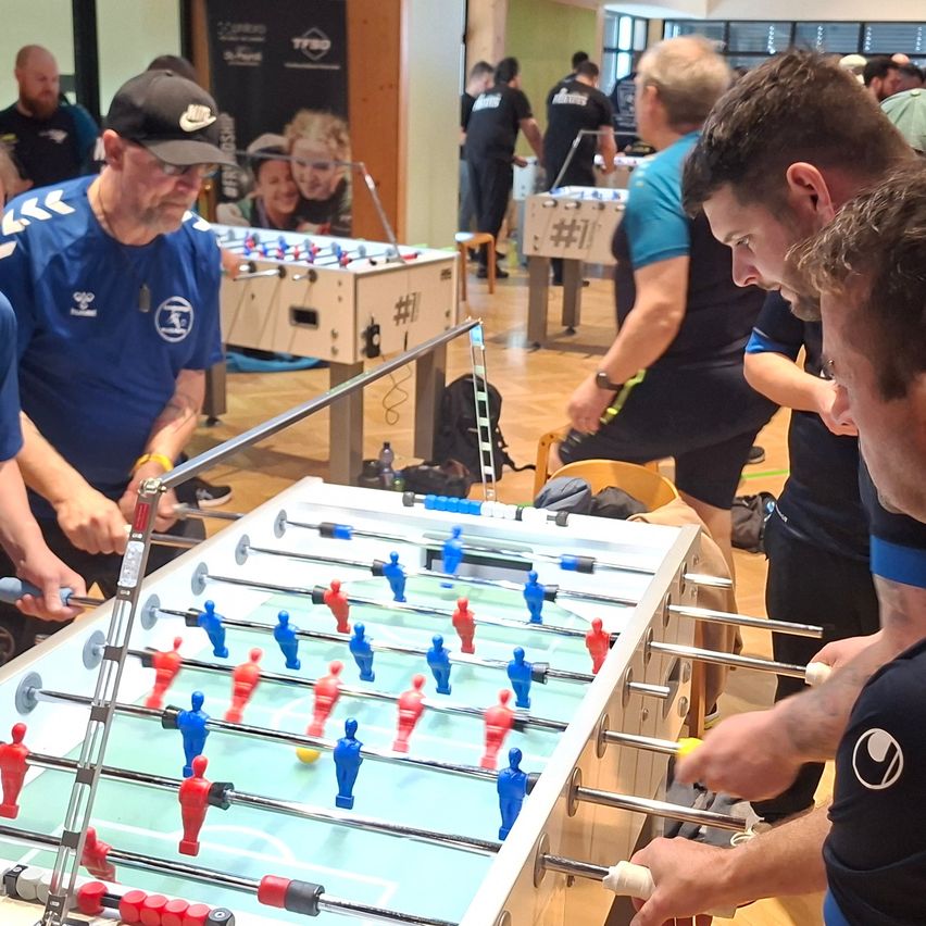A group of men play table football in a room. One man wears a blue shirt with a logo. Another wears a hat. A yellow ball rolls on the table.