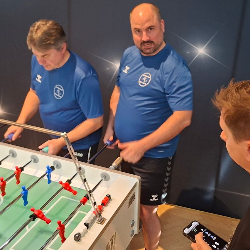 Two men in blue shirts with white logos play table football while a boy watches. A person holds a phone.