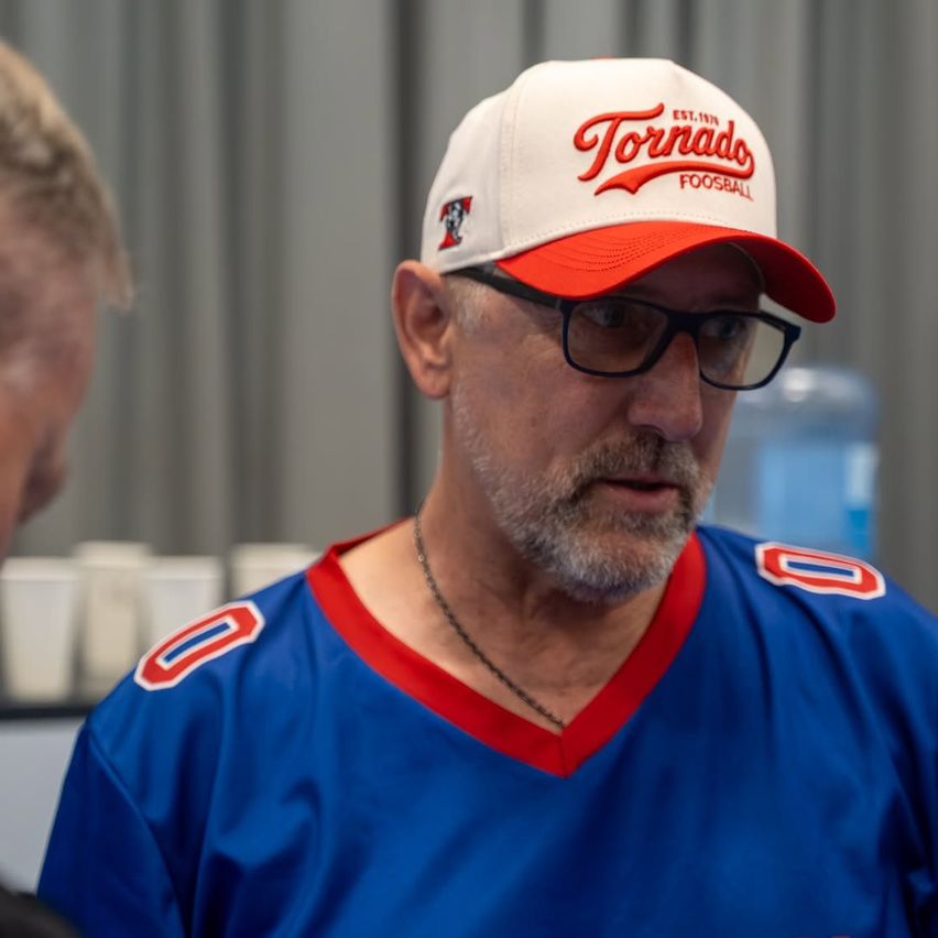 A man wearing a blue jersey with the number 0 and a Tornado football cap is conversing with someone in a room with cups and a water dispenser.