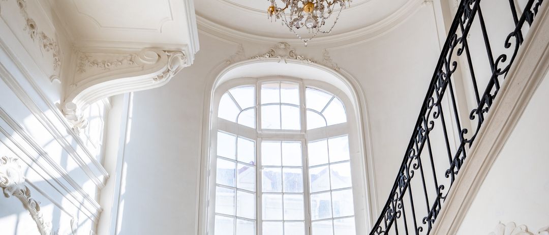 Eine große Treppe mit weißen Marmorstufen und einem schwarzen Eisengeländer. Die Treppe führt zu einem bogenförmigen Fenster mit weißen Rahmen.