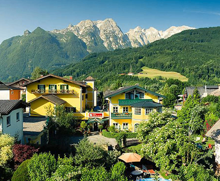 Luftaufnahme eines Dorfes mit gelben Häusern, grünen Bäumen und Bergen im Hintergrund unter einem blauen Himmel.
