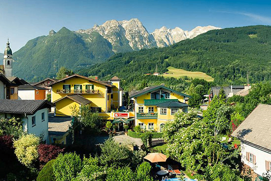 Luftaufnahme eines Dorfes mit gelben Häusern, grünen Bäumen und Bergen im Hintergrund unter einem blauen Himmel.