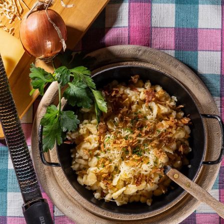 Ein Holzteller hält eine Schüssel mit cremigem Makkaroni-Käse, garniert mit knusprigen Zwiebeln und Käse. Eine Zwiebel und ein Messer liegen in der Nähe.