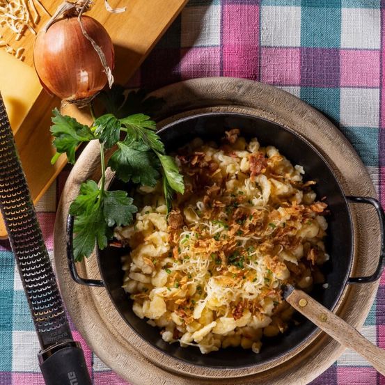 Ein Holzteller hält eine Schüssel mit cremigem Makkaroni-Käse, garniert mit knusprigen Zwiebeln und Käse. Eine Zwiebel und ein Messer liegen in der Nähe.