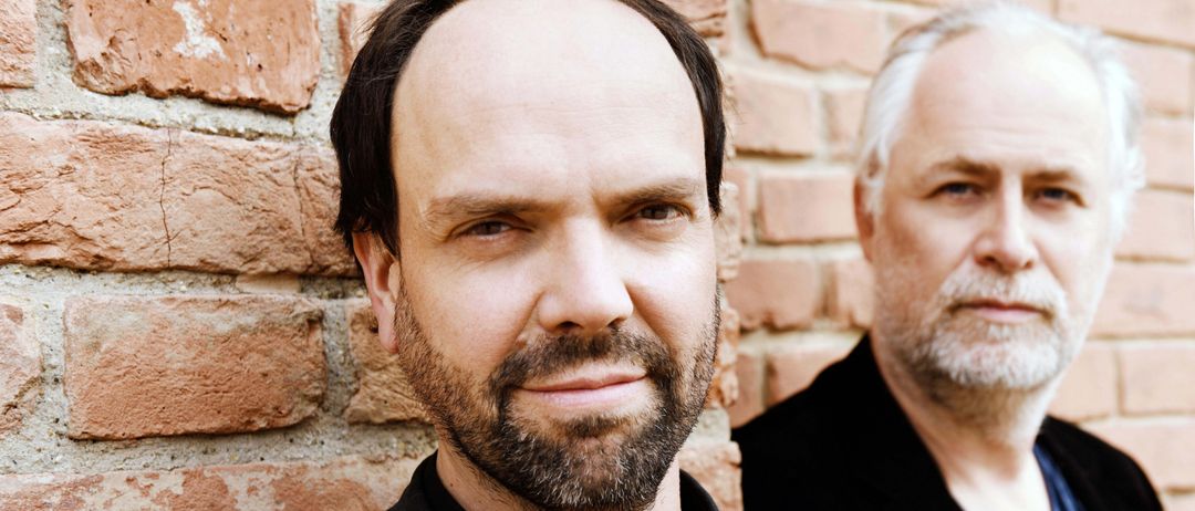 Zwei Männer stehen neben einer Ziegelmauer. Der Mann vorn hat einen Bart und trägt ein schwarzes Hemd. Der Mann dahinter hat weiße Haare und trägt einen schwarzen Mantel.