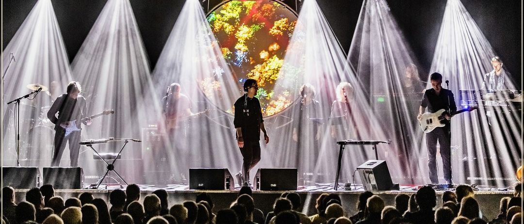 Eine Band tritt auf einer Bühne unter hellen Lichtern auf, mit einer kreisförmigen Projektion dahinter. Das Publikum sitzt davor und schaut die Show.