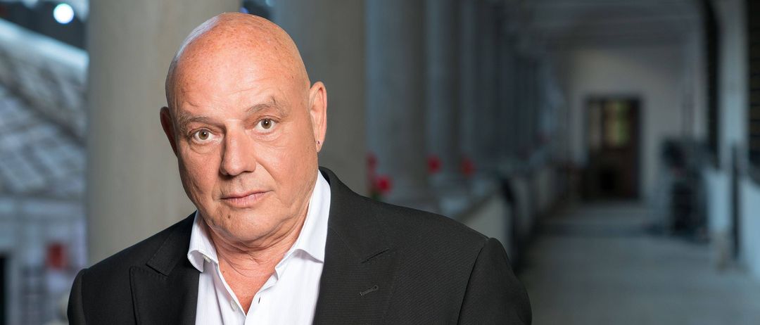 Ein kahlköpfiger Mann in einem schwarzen Anzug und weißem Hemd steht in einem Korridor und sieht geradeaus. Hinter ihm befinden sich weiße Säulen und rote Blumen.