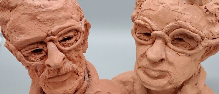 Zwei Tonfiguren eines älteren Mannes und einer Frau, beide mit Brille, sind vor einem grauen Hintergrund ausgestellt. Der Mann hat einen Schnurrbart und die Frau lächelt.