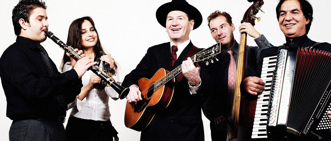 Eine Gruppe von Musikern, gekleidet in Anzügen, posiert für ein Foto. Ein Mann in der Mitte spielt Gitarre, während andere eine Klarinette, ein Akkordeon und eine Geige halten. Sie alle lächeln, mit einer weißen Wand als Hintergrund.