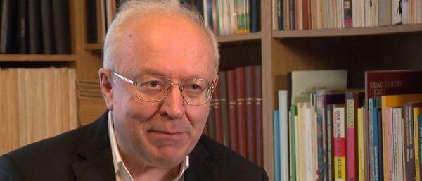 Ein älterer Mann mit Brille sitzt vor einem Bücherregal voller Bücher.