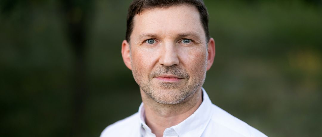 Ein Mann mit kurzem Bart und Schnurrbart in einem weißen Hemd posiert für ein Bild im Wald.