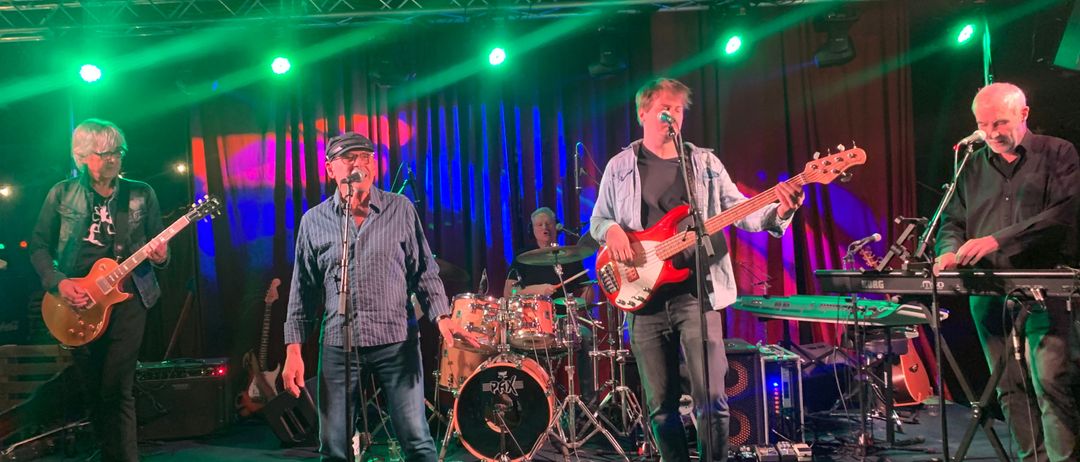 Eine Band spielt auf der Bühne unter grünen und roten Lichtern. Ein Mann singt, während ein anderer den Bass spielt. Hinter ihnen spielt ein weiterer Mann Schlagzeug. Die Bühne ist mit Musikinstrumenten gefüllt.