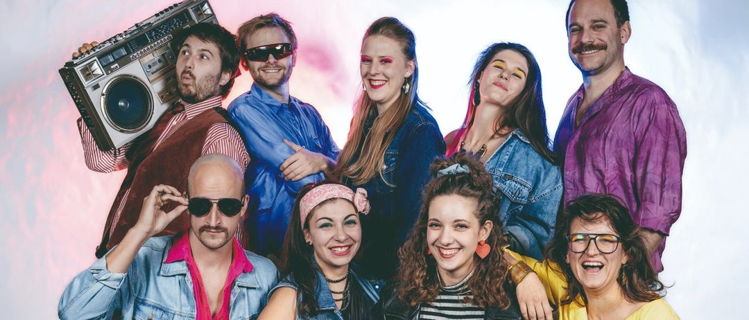 Eine Gruppe von Menschen in 1980er-Jahre-Kleidung posiert für ein Foto. Ein Mann hält eine Boombox, während andere Sonnenbrillen und Stirnbänder tragen.