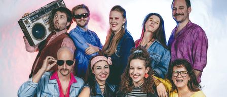 Eine Gruppe von Menschen in 1980er-Jahre-Kleidung posiert für ein Foto. Ein Mann hält eine Boombox, während andere Sonnenbrillen und Stirnbänder tragen.