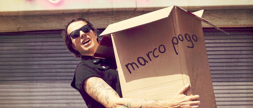 Ein Mann mit Sonnenbrille hält einen Karton mit der Aufschrift 'Marco Pogo' vor einem Neonschild mit der Aufschrift 'Verloren & Gefunden'. Er scheint für ein Foto zu posieren.