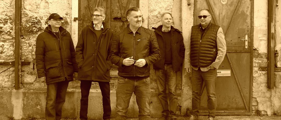 Fünf Männer stehen vor einem Gebäude mit alten Holztüren. Sie sind in Winterkleidung gekleidet. Sie lächeln und scheinen für ein Foto zu posieren.