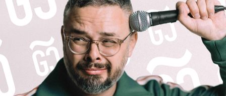 Ein Mann mit Brille und grüner Jacke hält ein Mikrofon in der rechten Hand, möglicherweise singend. Das Mikrofon ist schwarz mit einer silbernen Masche. Der Hintergrund hat Buchstaben und Symbole.