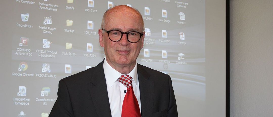 Ein Mann in einem schwarzen Anzug mit roter Krawatte und Brille steht vor einem Bildschirm mit einem Projektor.