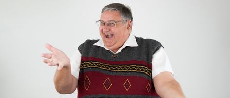 A man with gray hair is laughing, wearing glasses, a white shirt, and a patterned vest. He stands with arms outstretched against a plain white background.