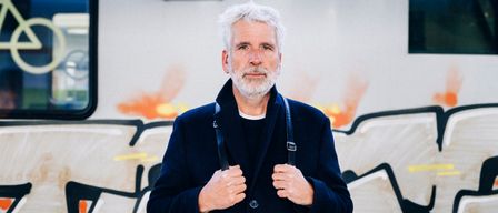 Ein älterer Mann mit weißem Bart und Schnurrbart steht vor einem mit Graffiti besprühten Zug. Er trägt einen dunklen Mantel mit einem weißen Innenshirt und hält die Schulterriemen seines Mantels.