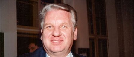 A smiling older man with gray hair and a mustache wearing a blue suit and tie poses for the camera.