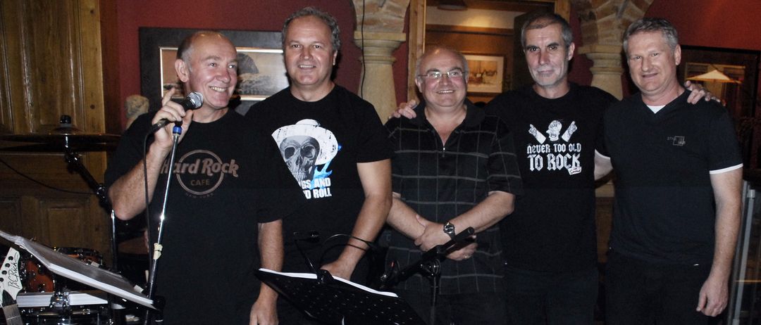 Fünf Männer posieren für ein Foto mit Musikinstrumenten in einem Raum mit roten Wänden und Ziegelbögen. Ein Mann hält ein Mikrofon.