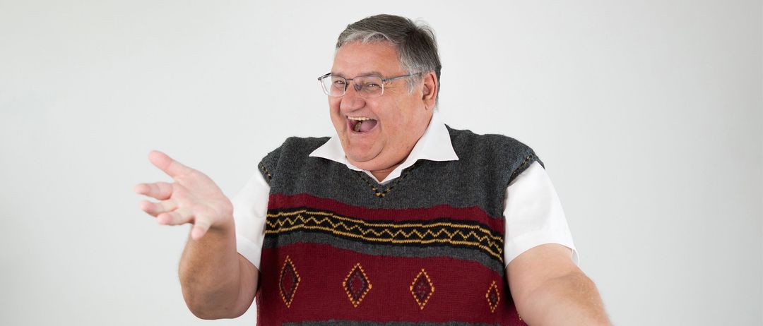 Ein Mann mit grauen Haaren und Brille steht mit offenem Mund und weit ausgestreckten Armen, trägt eine gemusterte Weste und schwarze Hose.