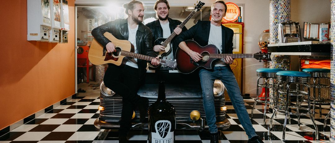 Drei Männer sitzen auf einem Oldtimer, halten Gitarren und haben eine Flasche vor sich, in einem Raum mit gemustertem Boden und Altstil-Dekor.