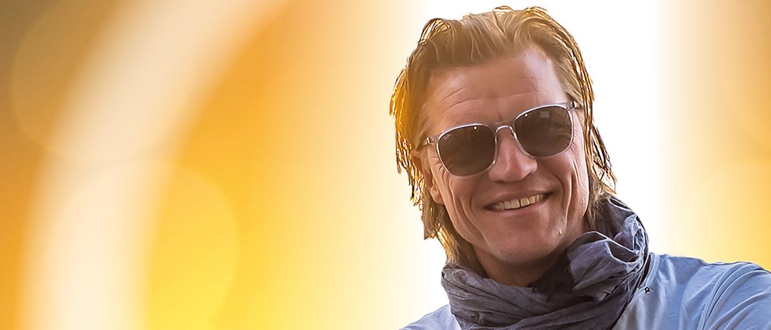 Ein Mann steht draußen, lächelt, trägt eine Sonnenbrille und einen Schal und hat die Arme verschränkt.