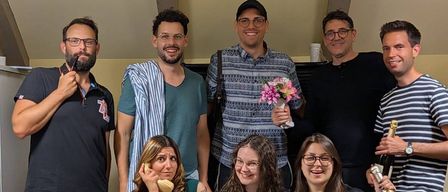 Eine Gruppe von Menschen posiert für ein Foto, wobei eine Person einen Blumenstrauß hält und eine andere eine Champagnerflasche. Eine Frau sitzt auf einem Stuhl, hält ein altes Telefon und lächelt. Ein Laptop steht auf einem Tisch vor ihnen und zeigt einen Videoanruf an.