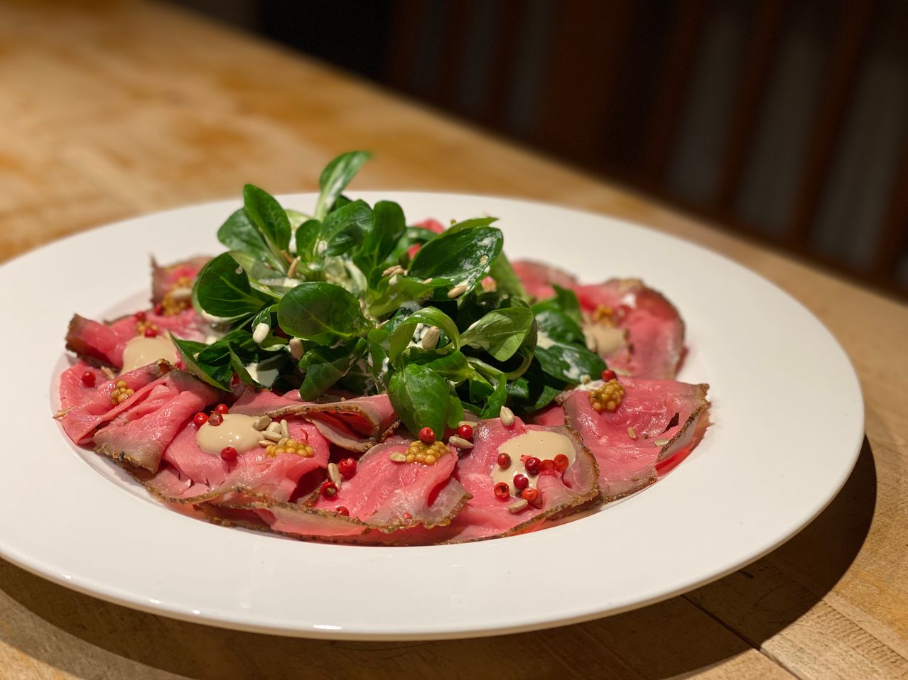 A white plate holds a dish of thinly sliced raw beef with a green salad, sprinkled with red and white toppings, placed on a wooden table.