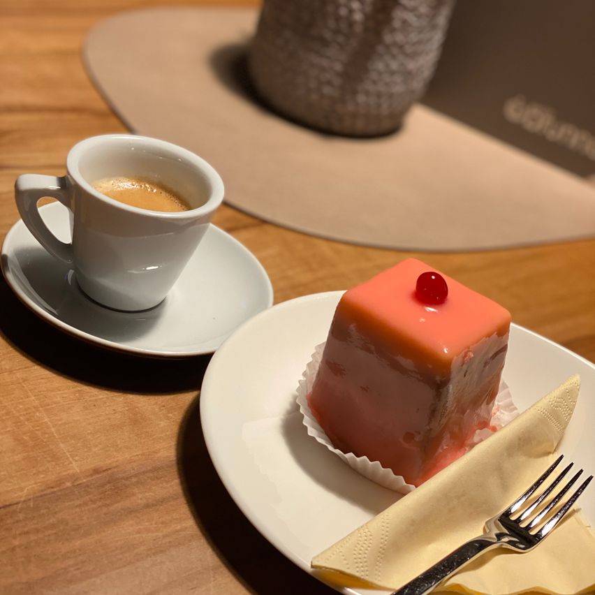 A cup of coffee with a saucer, and a dessert with a cherry on top are on a wooden table.